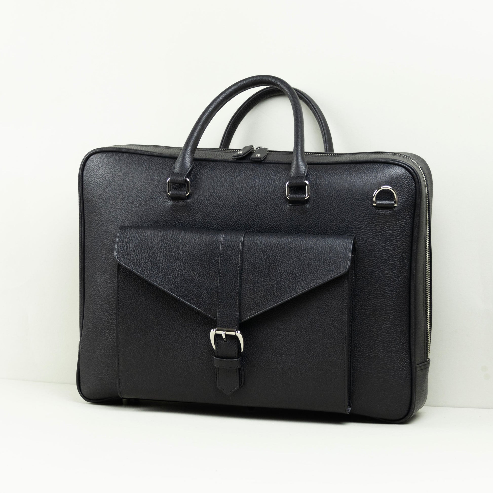 A black leather briefcase with dual top handles, a front flap pocket with a buckle closure, and silver-tone hardware, photographed on a white surface against a white wall.