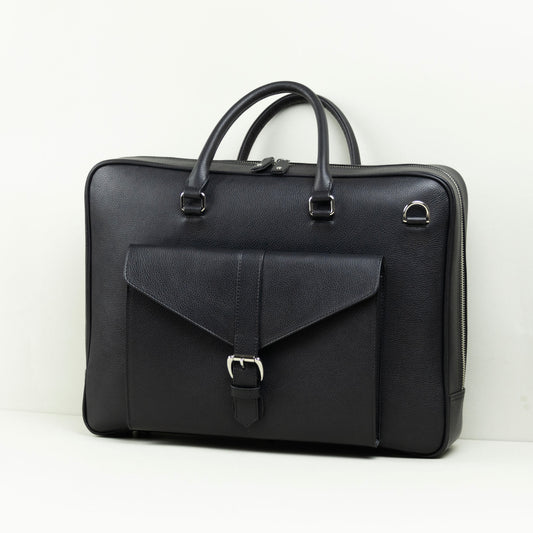 A black leather briefcase with dual top handles, a front flap pocket with a buckle closure, and silver-tone hardware, photographed on a white surface against a white wall.