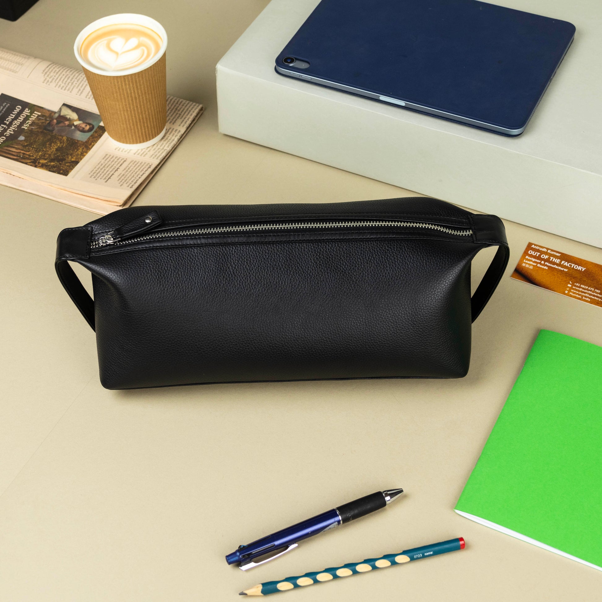 Black leather dopp kit on a desk with a cup, tablet, and stationery items.