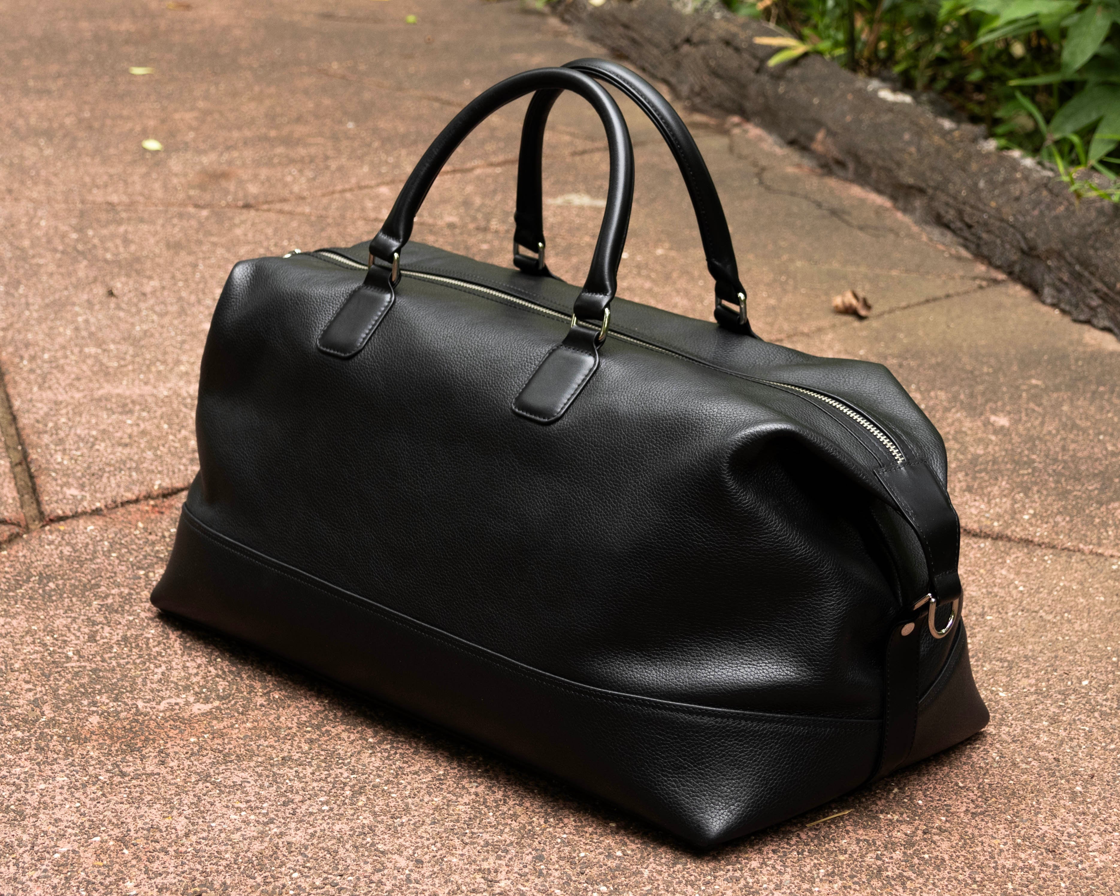 Black leather duffel bag on a concrete surface with greenery in the background